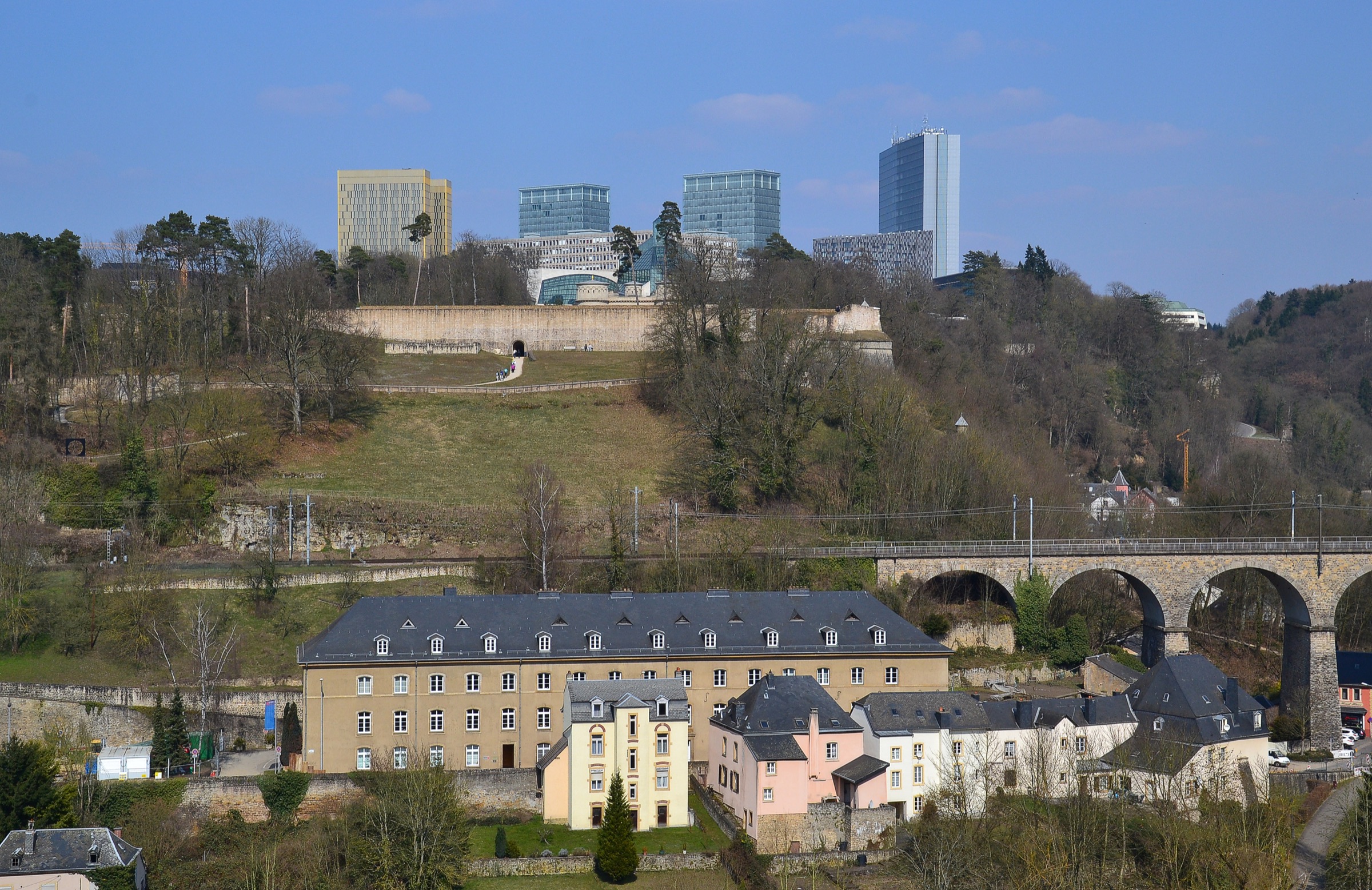 Sunny view of Luxembourg City's Kirchberg towers rising above the old city fortifications and the Clausen valley, seen under a clear blue sky.